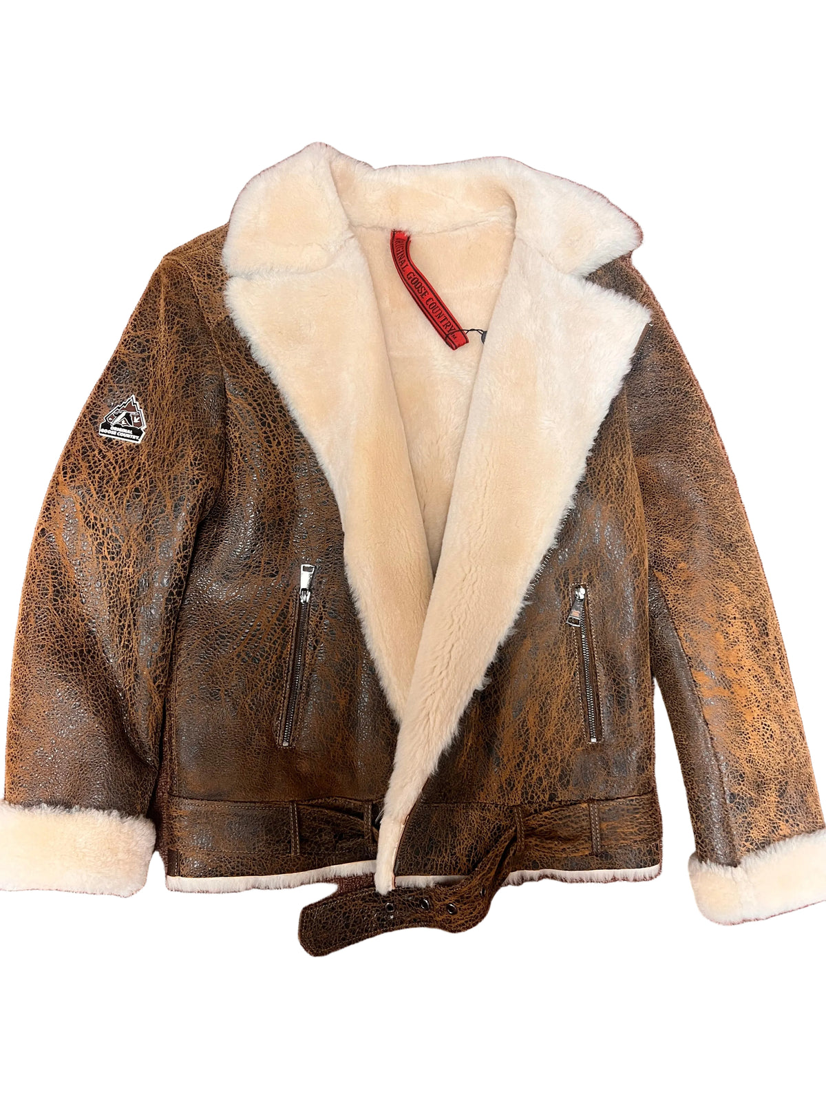 A brown distressed leather aviator jacket with a thick cream shearling lining, laid flat on a textured reddish-brown carpet. The jacket features a collar, zippered pockets, and a tie belt.