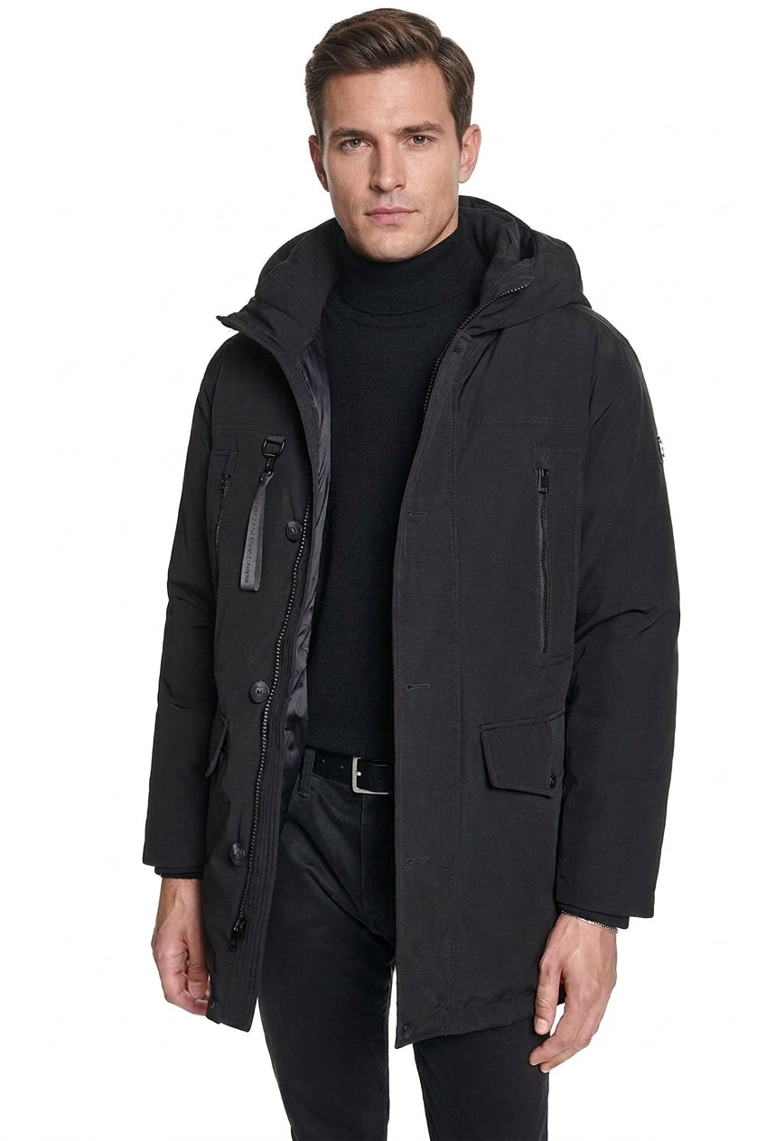 A man wearing a black hooded winter parka jacket over a black turtleneck sweater and dark pants, photographed against a plain white studio background.