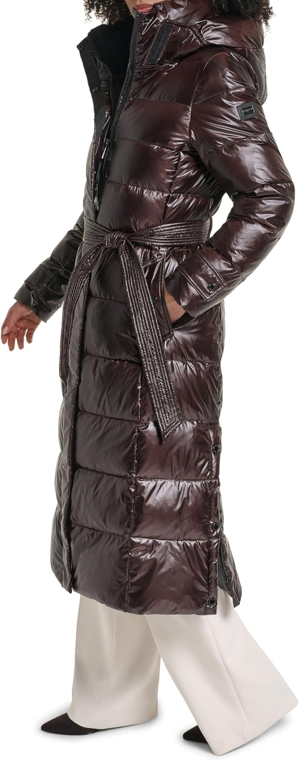 A long, glossy quilted puffer coat in a deep dark brown or burgundy tone, worn by a model with white wide-leg pants against a plain white background.