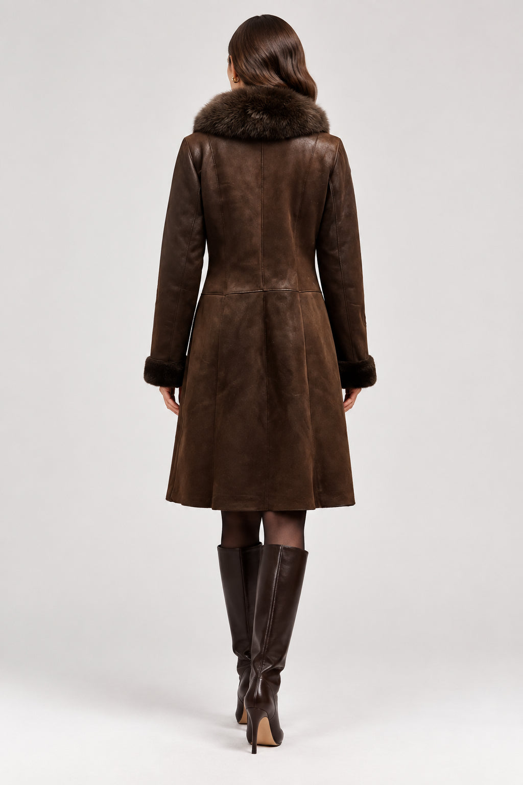 A dark brown suede shearling coat with a large dark brown fur collar, shown from the back on a dress form stand in a workshop setting.
