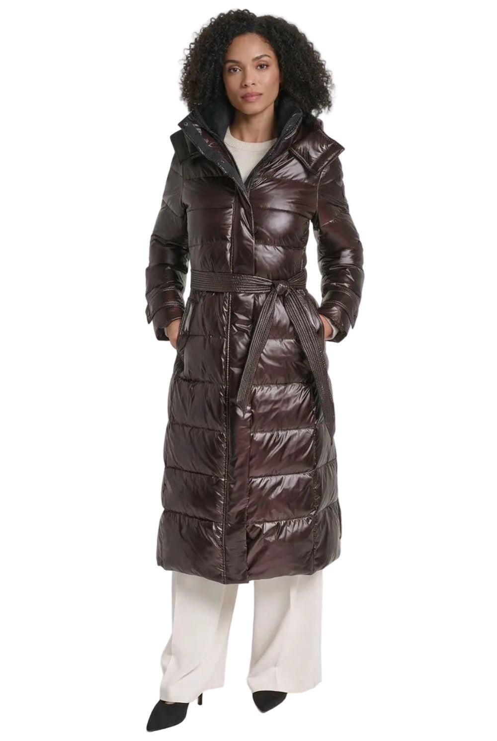 A woman wearing a long, glossy quilted puffer coat in dark brown with a matching belt, styled with cream wide-leg pants and dark pointed shoes.