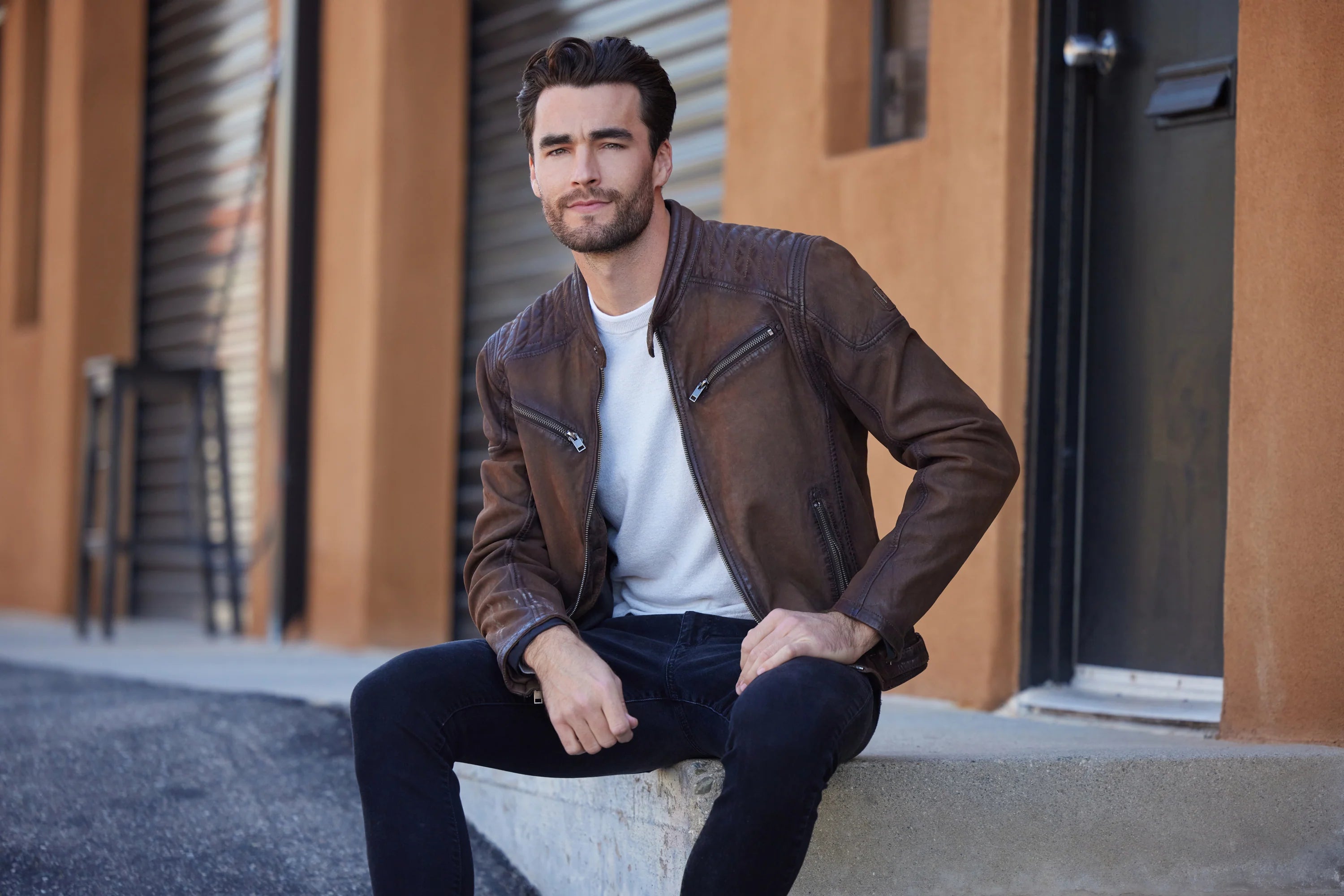 Man sitting on a concrete block wearing a brown leather jacket and dark pants.