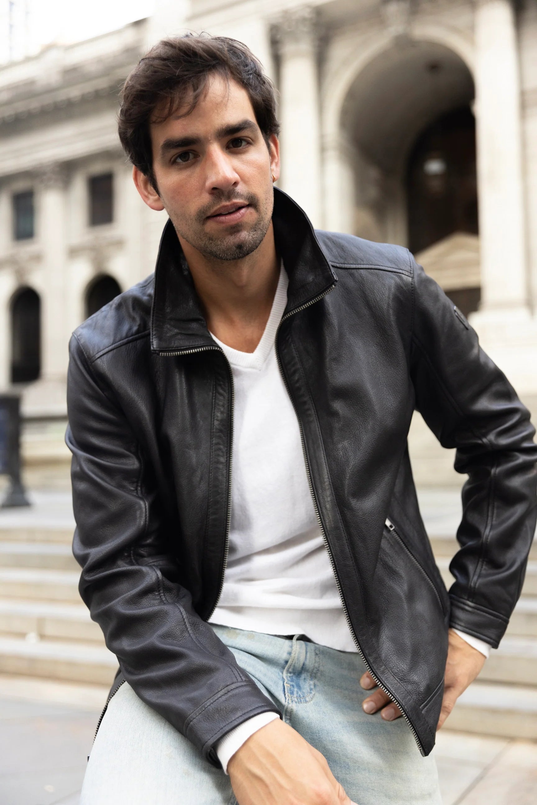 Man wearing a black leather jacket and light blue jeans sitting outdoors.