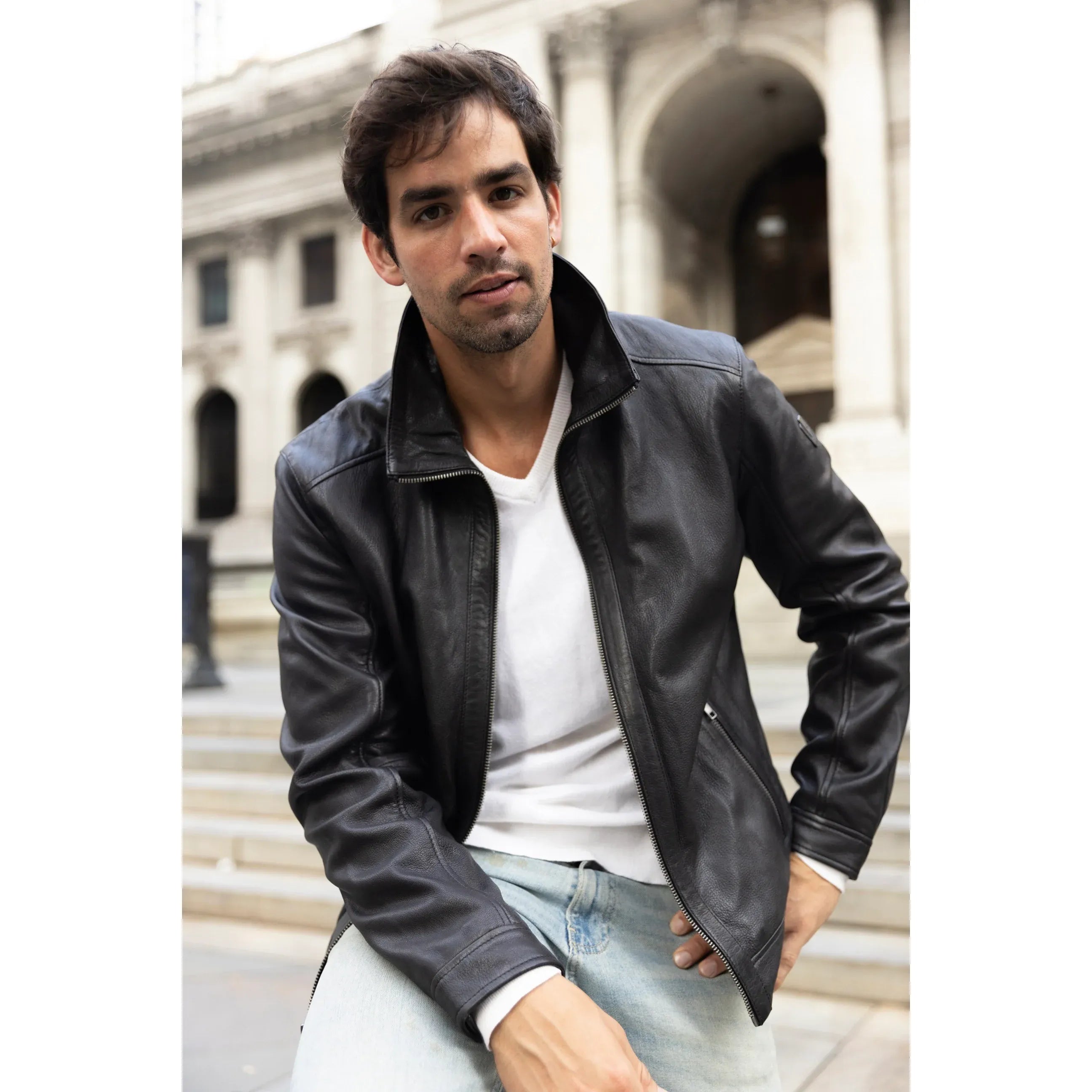 Man wearing a black leather jacket and light blue jeans sitting outdoors.